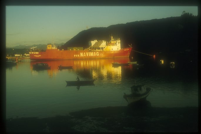 A large red ship docked by a hillside at sunset, reflecting on calm waters.